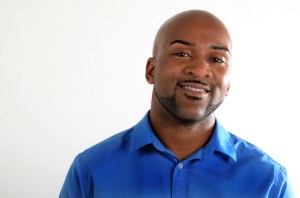 Headshot of Drew Anderson, a handsome Black man with a trimmed beard clean shaved head. He is wearing a royal blue button-up shirt and looking directly into the camera.