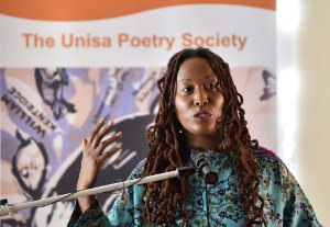 Action photo of Natalia Molebatsi speaking in front of a mic with her hand raised slightly gesticulating. She's wearing a light green top and her locs are draping down over her shoulders. A sign behind her says The Unisa Poetry Society.